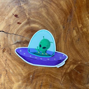 May include: A green alien with large eyes is sitting in a purple and blue spaceship with a clear dome. The spaceship has a white outline and small white dots around the edge. The sticker has the text "@kait.k.art" in the bottom right corner.