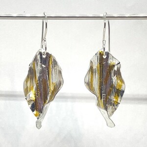 May include: A pair of silver earrings with a textured, leaf-shaped design. The leaves are accented with gold and have a slightly wavy appearance.