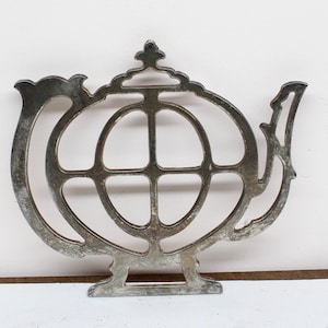 Puede incluir: Calientaplatos de metal plateado en forma de tetera con un diseño circular en el centro. El calientaplatos tiene una parte superior decorativa y una base con tres patas.