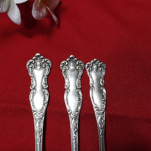 May include: Three ornate silver spoons with detailed floral and scrollwork designs are arranged against a deep red background. The spoons have a classic, vintage aesthetic, with a decorative handle and a polished finish. White flowers are in the upper left corner.