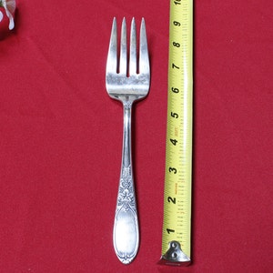 May include: A silver fork with an ornate design on the handle. The fork is 7 inches long and is measured against a yellow tape measure.