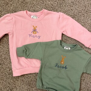 May include: Two children's sweatshirts. One is pink with the name "Mary" embroidered below a bunny design. The other is green with the name "Jacob" embroidered below a bunny design. Both have long sleeves and are made of a soft material.