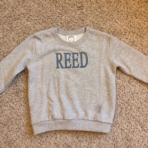 Könnte beinhalten: Ein graues Sweatshirt mit dem Namen "REED" in marineblauen Blockbuchstaben auf der Brust gestickt. Das Sweatshirt hat einen Rundhalsausschnitt und lange Ärmel. Das Kleidungsstück ist aus einem weichen Material gefertigt.