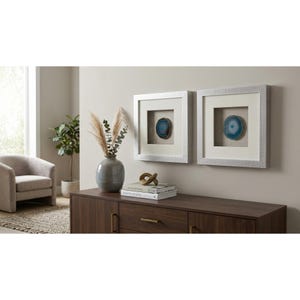 May include: Two square wall frames with silver textured borders, each containing a centered blue agate slice on a neutral fabric background. The frames are mounted side-by-side on a wall above a dark wood cabinet with gold-toned hardware.