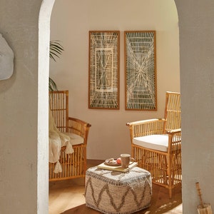 May include: Two wicker chairs with white cushions and a woven ottoman in a room with two framed geometric wall hangings.