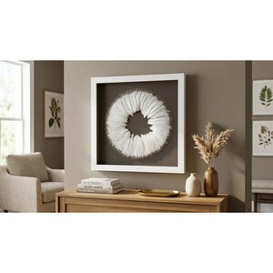 May include: A square white shadow box frame containing a circular arrangement of white feathers against a dark gray background. The artwork is displayed on a wooden surface alongside a small gold tray, a white vase, and a brown vase with dried grass.
