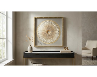 May include: A square wall art piece featuring a circular, pleated, white and gold sunburst design encased in a gold frame. The artwork is mounted on a wall above a dark console table with a white top, a ceramic vase, and stacked books.