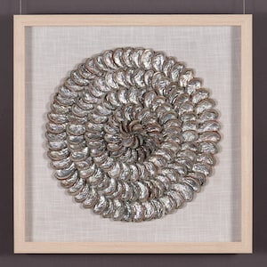 May include: Circular wall art featuring a spiral design made of silver-colored, shell-like objects. The artwork is set against a white fabric background and framed in a light-colored wooden frame. The overall aesthetic is coastal and decorative.