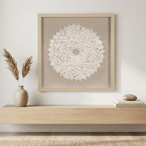 May include: A square wall art piece featuring a circular arrangement of white seashells mounted on a beige fabric background within a light wood frame. The artwork is displayed above a long, horizontal light wood floating shelf.