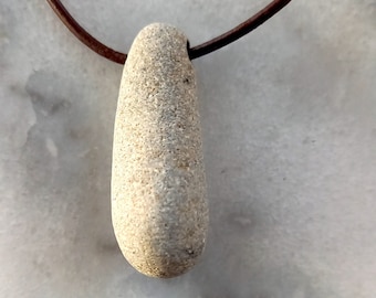 May include: A light gray stone pendant on a brown leather cord. The stone is smooth and has a slightly rounded shape.