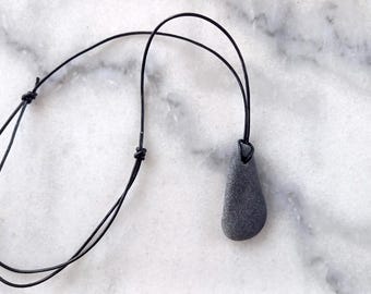 May include: A black leather cord necklace with a gray stone pendant. The stone is smooth and has a teardrop shape.