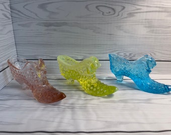 May include: Three glass slipper-shaped dishes in pink, yellow, and blue. The dishes are decorated with raised patterns and have a vintage look.