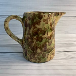 May include: A small, brown and green ceramic pitcher with a speckled glaze. The pitcher has a handle and a spout.