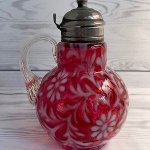 May include: A vintage glass creamer or sugar dispenser with a silver lid. The glass is a deep red colour with a white floral pattern.