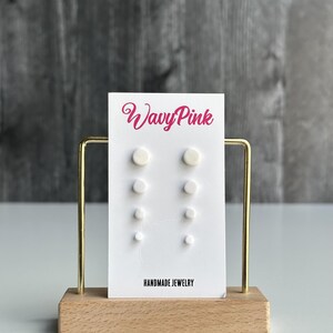 May include: A set of four pairs of white circular stud earrings displayed on a white card with the text WavyPink and HANDMADE JEWELRY. The card is held by a gold metal frame attached to a rectangular wooden base, presenting a clean jewelry display.