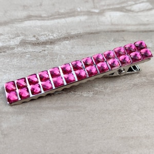 May include: A rectangular hair clip with two rows of bright pink rhinestones. The clip is made of silver-colored metal and has a hinged design for securing hair. The rhinestones are square-cut and closely set.