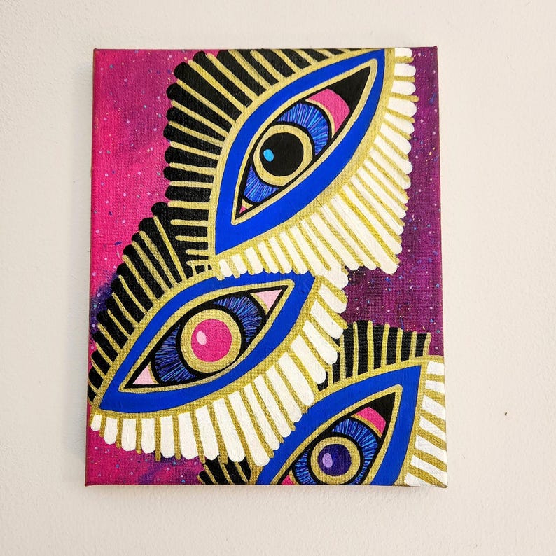 May include: An abstract painting on canvas featuring three stylized eye designs. Each eye has a blue iris, pink center, and gold and white feather-like accents against a pink and purple speckled background. The artwork is framed.