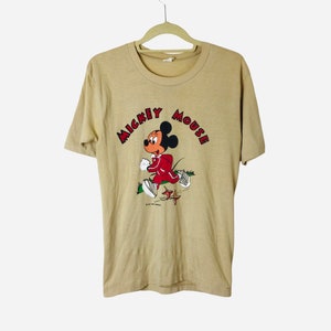 K&ouml;nnte beinhalten: Beiges T-Shirt mit einem laufenden Mickey Mouse-Motiv. Mickey tr&auml;gt einen roten Trainingsanzug und wei&szlig;e Turnschuhe. Der Schriftzug "Mickey Mouse" ist rot. Das T-Shirt h&auml;ngt an einem beigen Kleiderb&uuml;gel.