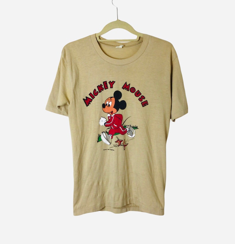K&ouml;nnte beinhalten: Beiges T-Shirt mit einem laufenden Mickey Mouse-Motiv. Mickey tr&auml;gt einen roten Trainingsanzug und wei&szlig;e Turnschuhe. Der Schriftzug "Mickey Mouse" ist rot. Das T-Shirt h&auml;ngt an einem beigen Kleiderb&uuml;gel.
