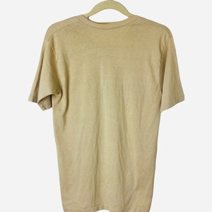 K&ouml;nnte beinhalten: Ein hellbraunes, kurz&auml;rmeliges T-Shirt h&auml;ngt an einem beigen Kleiderb&uuml;gel vor wei&szlig;em Hintergrund. Das Shirt besteht aus einem weichen, schlichten Stoff. Ein einfaches, l&auml;ssiges Kleidungsst&uuml;ck.