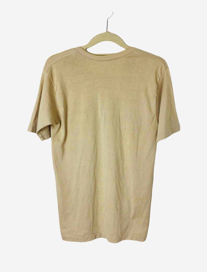 K&ouml;nnte beinhalten: Ein hellbraunes, kurz&auml;rmeliges T-Shirt h&auml;ngt an einem beigen Kleiderb&uuml;gel vor wei&szlig;em Hintergrund. Das Shirt besteht aus einem weichen, schlichten Stoff. Ein einfaches, l&auml;ssiges Kleidungsst&uuml;ck.