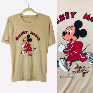 K&ouml;nnte beinhalten: Beiges T-Shirt mit einer Grafik von Mickey Mouse in einem roten Trainingsanzug, der rennt. Der Text "Mickey Mouse" ist in roter Farbe &uuml;ber der Figur angeordnet. Das T-Shirt h&auml;ngt an einem Holzb&uuml;gel.