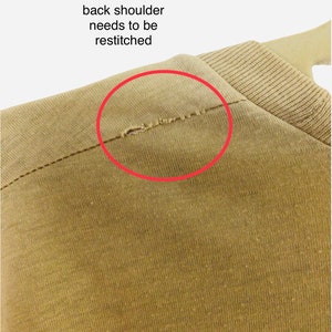 K&ouml;nnte beinhalten: Nahaufnahme eines beigefarbenen Kleidungsst&uuml;cks, mit einem kleinen Bereich an der hinteren Schulter, der neu vern&auml;ht werden muss, markiert durch einen roten Kreis. Der Stoff scheint ein Strickmaterial zu sein, und das Kleidungsst&uuml;ck h&auml;ngt an einem Kleiderb&uuml;gel.