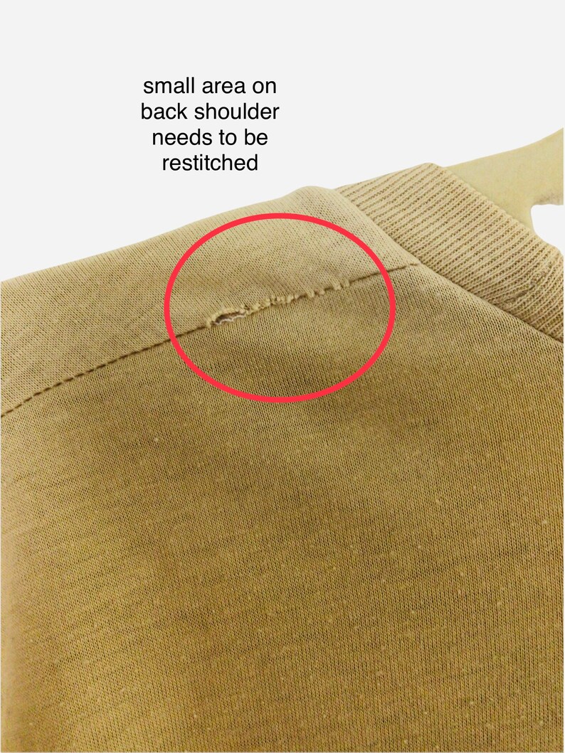 K&ouml;nnte beinhalten: Nahaufnahme eines beigefarbenen Kleidungsst&uuml;cks, mit einem kleinen Bereich an der hinteren Schulter, der neu vern&auml;ht werden muss, markiert durch einen roten Kreis. Der Stoff scheint ein Strickmaterial zu sein, und das Kleidungsst&uuml;ck h&auml;ngt an einem Kleiderb&uuml;gel.