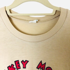 K&ouml;nnte beinhalten: Beigefarbenes T-Shirt mit Rundhalsausschnitt und den Worten "Mickey Mouse" in Rot und Schwarz. Das Shirt h&auml;ngt an einem beigen Kleiderb&uuml;gel. Das Bild ist eine Nahaufnahme des oberen Teils des Shirts.