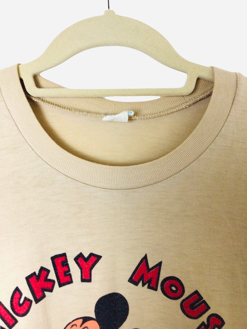 K&ouml;nnte beinhalten: Beigefarbenes T-Shirt mit Rundhalsausschnitt und den Worten "Mickey Mouse" in Rot und Schwarz. Das Shirt h&auml;ngt an einem beigen Kleiderb&uuml;gel. Das Bild ist eine Nahaufnahme des oberen Teils des Shirts.