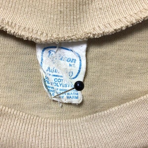 K&ouml;nnte beinhalten: Nahaufnahme eines beigefarbenen T-Shirt-Kragens mit einem eingen&auml;hten Etikett. Das Etikett ist wei&szlig; mit blauem Text und einer schwarzen Nadel. Das Etikett lautet "50% COTTON, 50% POLYESTER" und "MASCHINENW&Auml;SCHE WARM, TROCKNER GEEIGNET."