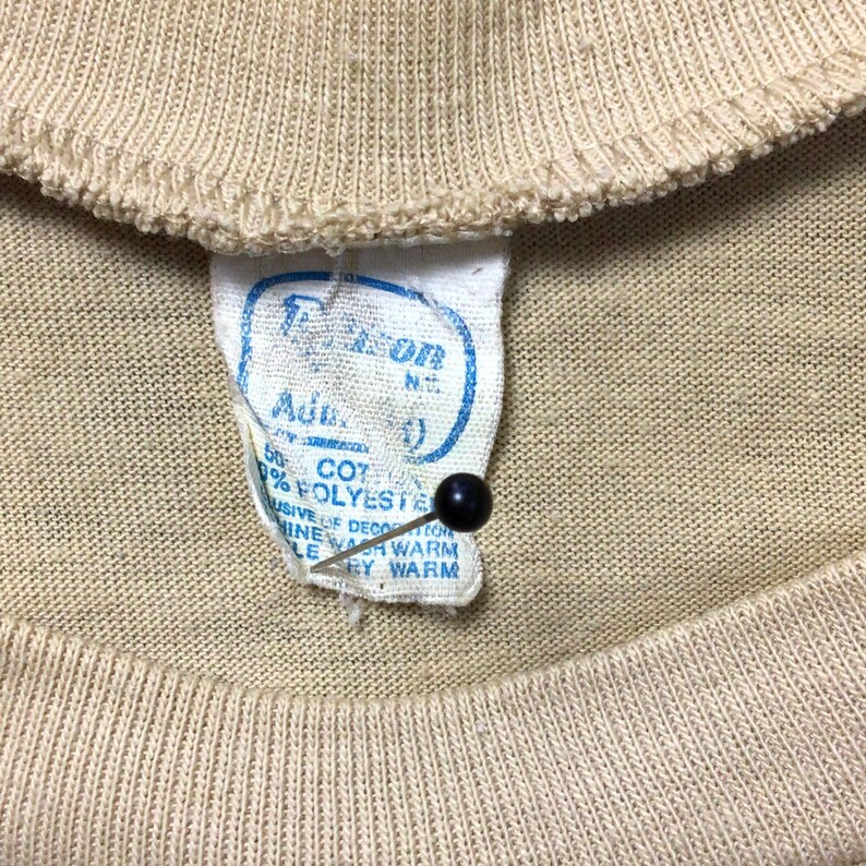 K&ouml;nnte beinhalten: Nahaufnahme eines beigefarbenen T-Shirt-Kragens mit einem eingen&auml;hten Etikett. Das Etikett ist wei&szlig; mit blauem Text und einer schwarzen Nadel. Das Etikett lautet "50% COTTON, 50% POLYESTER" und "MASCHINENW&Auml;SCHE WARM, TROCKNER GEEIGNET."