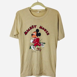 K&ouml;nnte beinhalten: Beigefarbenes T-Shirt mit einer Grafik von Mickey Mouse in einem roten Trainingsanzug, der rennt. Der Text "MICKEY MOUSE" ist in roten Buchstaben &uuml;ber der Figur angeordnet. Das Shirt h&auml;ngt an einem beigen Kleiderb&uuml;gel.
