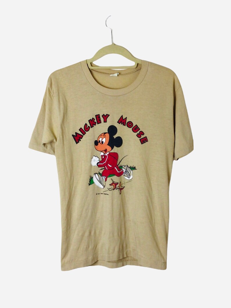 K&ouml;nnte beinhalten: Beigefarbenes T-Shirt mit einer Grafik von Mickey Mouse in einem roten Trainingsanzug, der rennt. Der Text "MICKEY MOUSE" ist in roten Buchstaben &uuml;ber der Figur angeordnet. Das Shirt h&auml;ngt an einem beigen Kleiderb&uuml;gel.