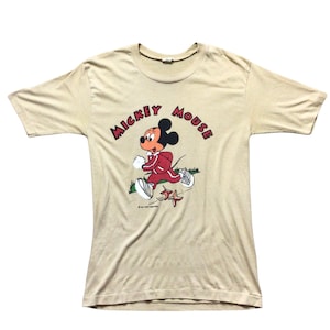 K&ouml;nnte beinhalten: Beigefarbenes T-Shirt mit einer Grafik von Mickey Mouse in einem roten Trainingsanzug, der rennt. Die Worte "Mickey Mouse" sind in roten Buchstaben &uuml;ber der Figur angeordnet. Das Shirt hat einen Vintage-Stil.