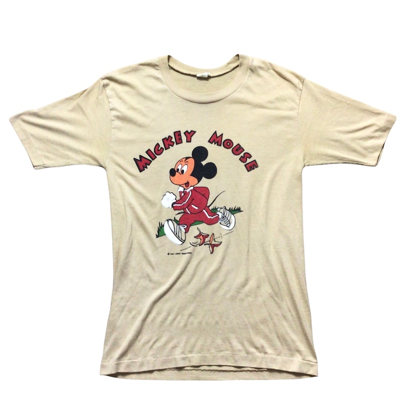 K&ouml;nnte beinhalten: Beigefarbenes T-Shirt mit einer Grafik von Mickey Mouse in einem roten Trainingsanzug, der rennt. Die Worte "Mickey Mouse" sind in roten Buchstaben &uuml;ber der Figur angeordnet. Das Shirt hat einen Vintage-Stil.