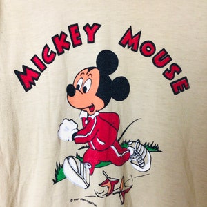 K&ouml;nnte beinhalten: Beiges T-Shirt mit einem laufenden Mickey Mouse-Motiv. Die Zeichentrickfigur tr&auml;gt einen roten Trainingsanzug und wei&szlig;e Turnschuhe. Die Worte "MICKEY MOUSE" sind in roten Buchstaben &uuml;ber der Figur angeordnet. Ein Vintage-Disney-Produkt.