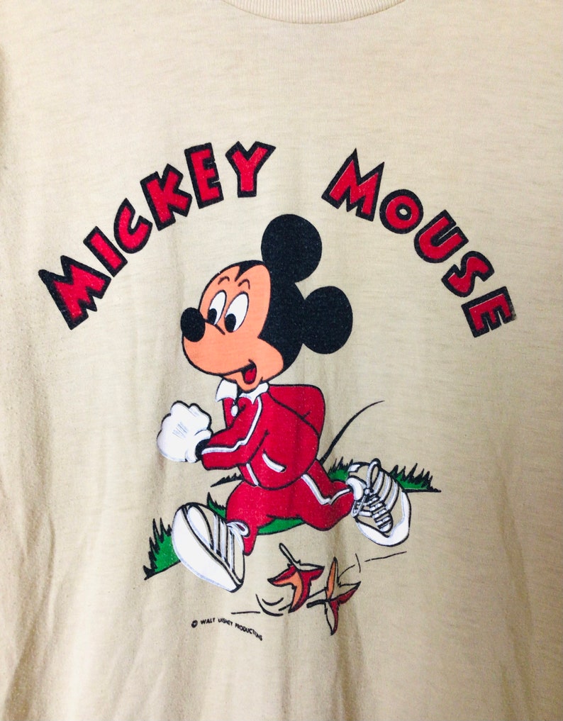 K&ouml;nnte beinhalten: Beiges T-Shirt mit einem laufenden Mickey Mouse-Motiv. Die Zeichentrickfigur tr&auml;gt einen roten Trainingsanzug und wei&szlig;e Turnschuhe. Die Worte "MICKEY MOUSE" sind in roten Buchstaben &uuml;ber der Figur angeordnet. Ein Vintage-Disney-Produkt.