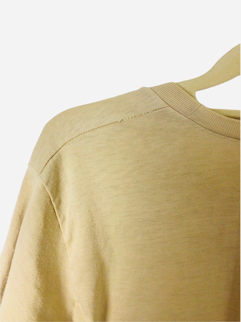 K&ouml;nnte beinhalten: Nahaufnahme eines hellbraunen Rundhals-T-Shirts. Die Schulternaht ist sichtbar, mit einer Nahtlinie. Das Shirt h&auml;ngt an einem Holzkleiderb&uuml;gel. Der Stoff hat ein leicht strukturiertes Aussehen.