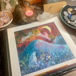 May include: A watercolor print of three white hares leaping over a hill with a crescent moon in the sky. The hares are silhouetted against a colorful background of blues, purples, and oranges. The print is framed and ready to hang.