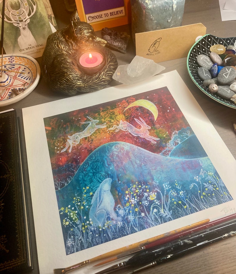 May include: A watercolor print of three white hares leaping over a hill with a crescent moon in the sky. The hares are silhouetted against a colorful background of blues, purples, and oranges. The print is framed and ready to hang.