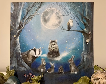 May include: A greeting card featuring a whimsical illustration of a cat, badger, rabbits, and an owl gathered under a full moon in a forest setting. The card has a blue and white color scheme with gold accents.