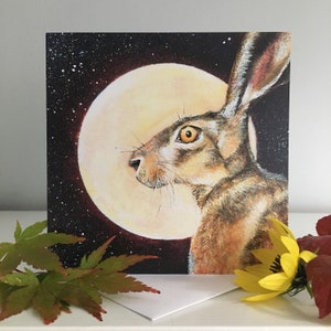 May include: A greeting card featuring a watercolor illustration of a brown hare looking up at a full moon against a dark blue starry night sky.
