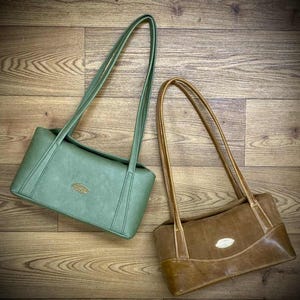 May include: Two shoulder bags, one sage green and one brown, are displayed from above. Both have long, thin straps and a rectangular shape. Each bag features a small, oval-shaped gold emblem on the front.