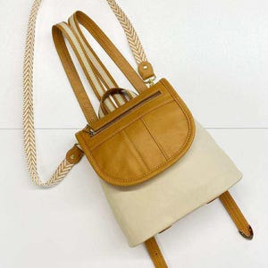 Puede incluir: Una mochila de color beige y crema con solapa de cuero y cuerpo crema. La mochila tiene correas de cuero marrón claro y una correa estampada de color crema. La bolsa tiene cierre de cremallera y herrajes metálicos.