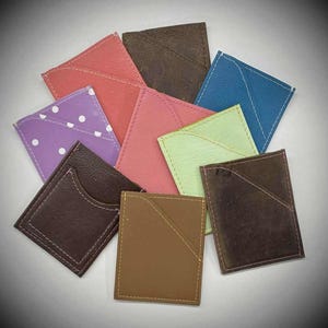 May include: A collection of colorful, small leather cardholders in various shades, including pink, brown, blue, and purple. Each cardholder has a pocket and is stitched with contrasting thread. The cardholders are arranged in a circular pattern.