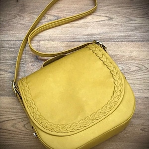 Puede incluir: Un bolso bandolera amarillo mostaza con un ribete trenzado alrededor de la solapa. El bolso tiene una correa larga y un cierre de solapa.