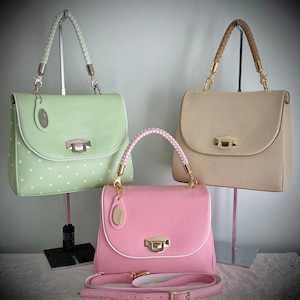 Könnte beinhalten: Drei unterschiedlich farbige Handtaschen mit weißem Besatz und goldenen Beschlägen. Die Handtaschen werden auf Ständern präsentiert. Die Handtaschen sind grün mit weißen Punkten, beige und rosa.