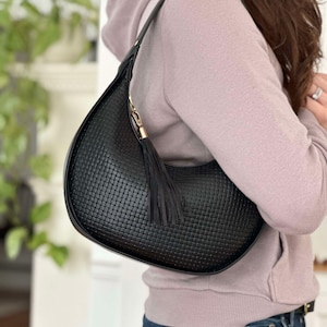 May include: Black woven leather shoulder bag with a curved shape. The bag features a long strap, a gold-tone clasp, and a black tassel. The bag is carried over the shoulder.