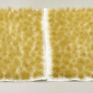 May include: Two rectangular sheets of model railroad scenery material. The sheets are covered in a light tan, grass-like texture, with a white border. The material is designed to simulate dry grass or a field for model landscapes.